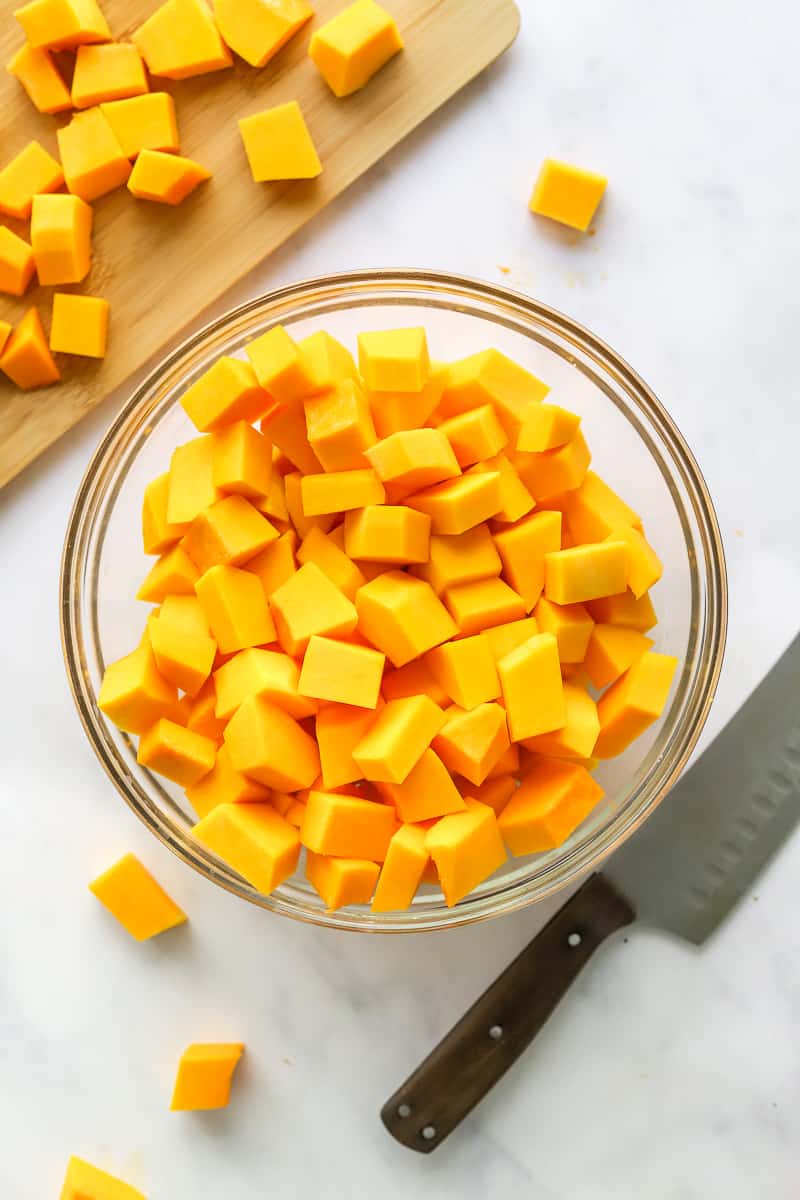 Round, glass bowl filled with cubed butternut squash with more cubes squash on a wooden cutting board behind it. 