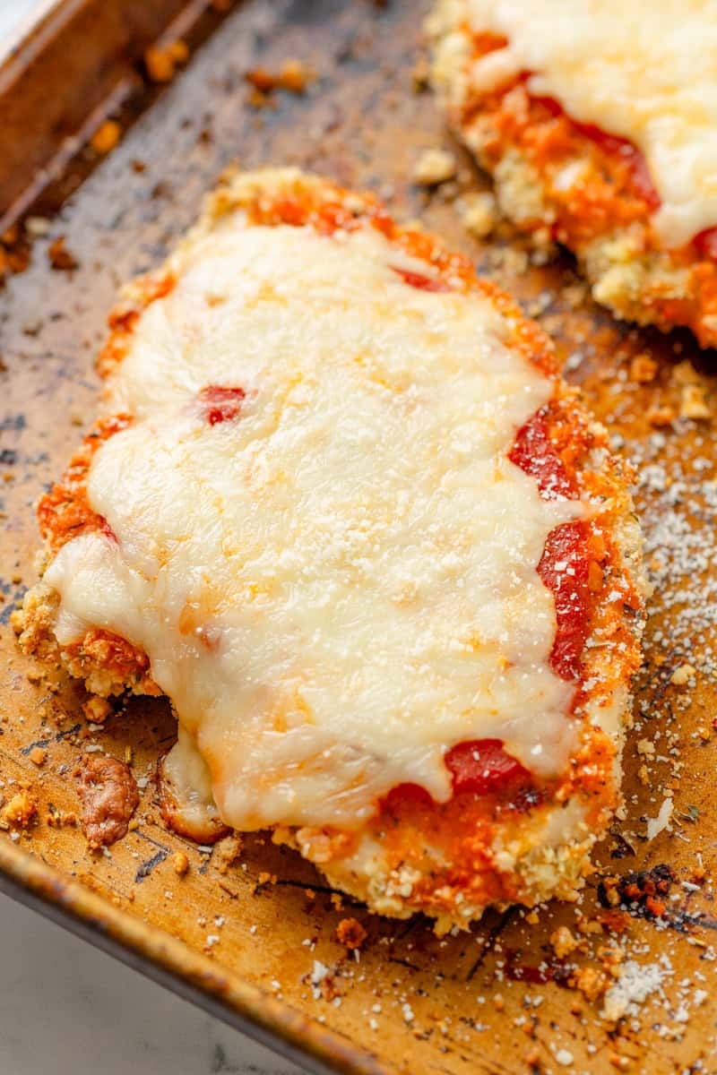 Golden cooked breaded chicken breast topped with sauce and melty cheese on a pan with more behind it. 