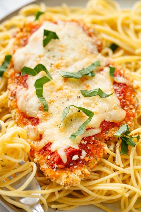Cooked gluten free chicken parmesan on a bed of cooked spaghetti with basil on top of it.
