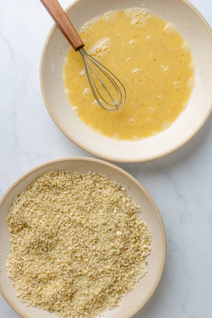 Bowl of breadcrumbs with a bowl of whisked eggs with a whisk in the bowl behind it. 