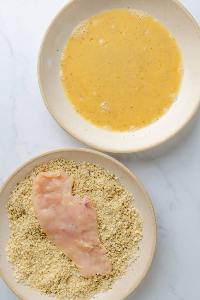 Raw chicken breast in a bowl of breadcrumbs with a bowl of whisked eggs behind it. 