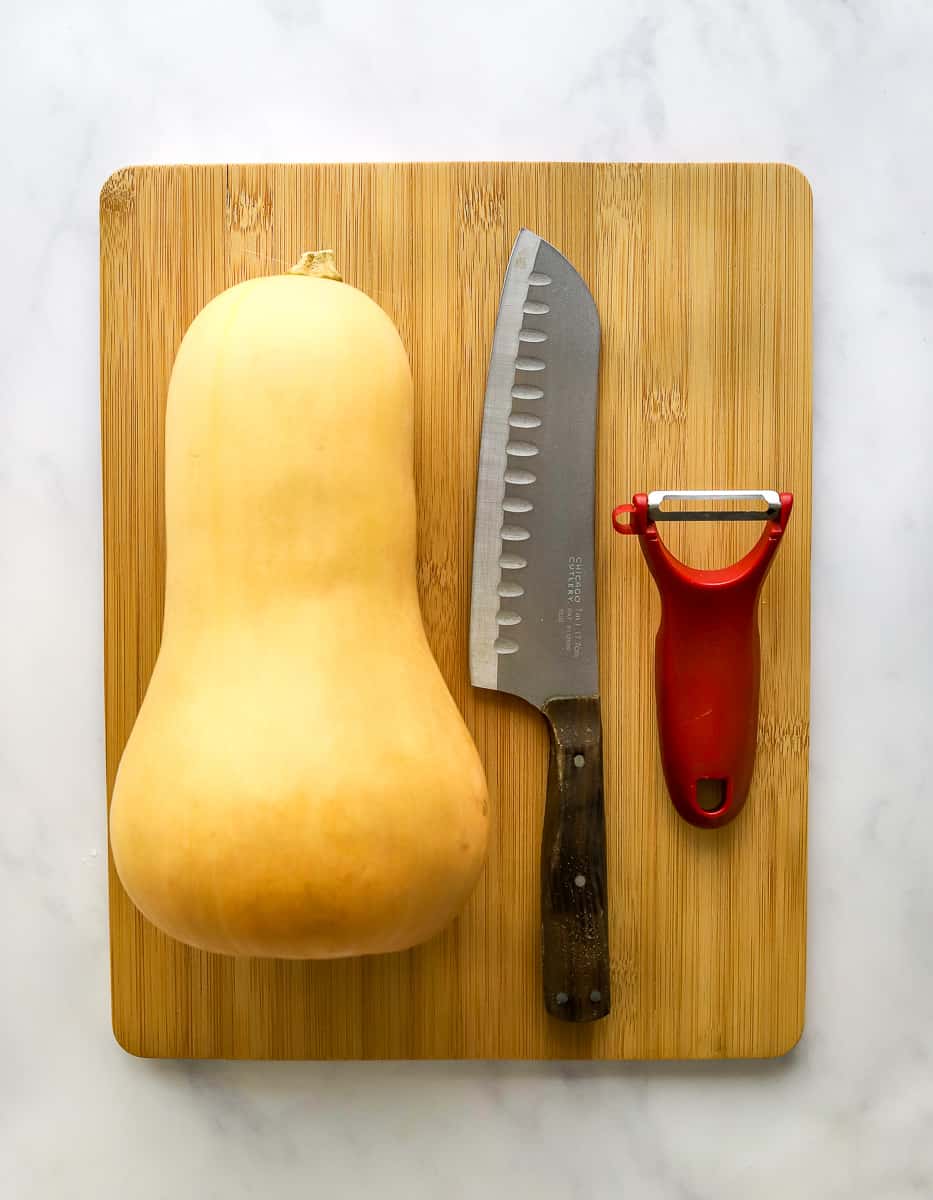 Whole butternut squash on a cutting board with a kitchen knife and a red vegetable peeler next to it.