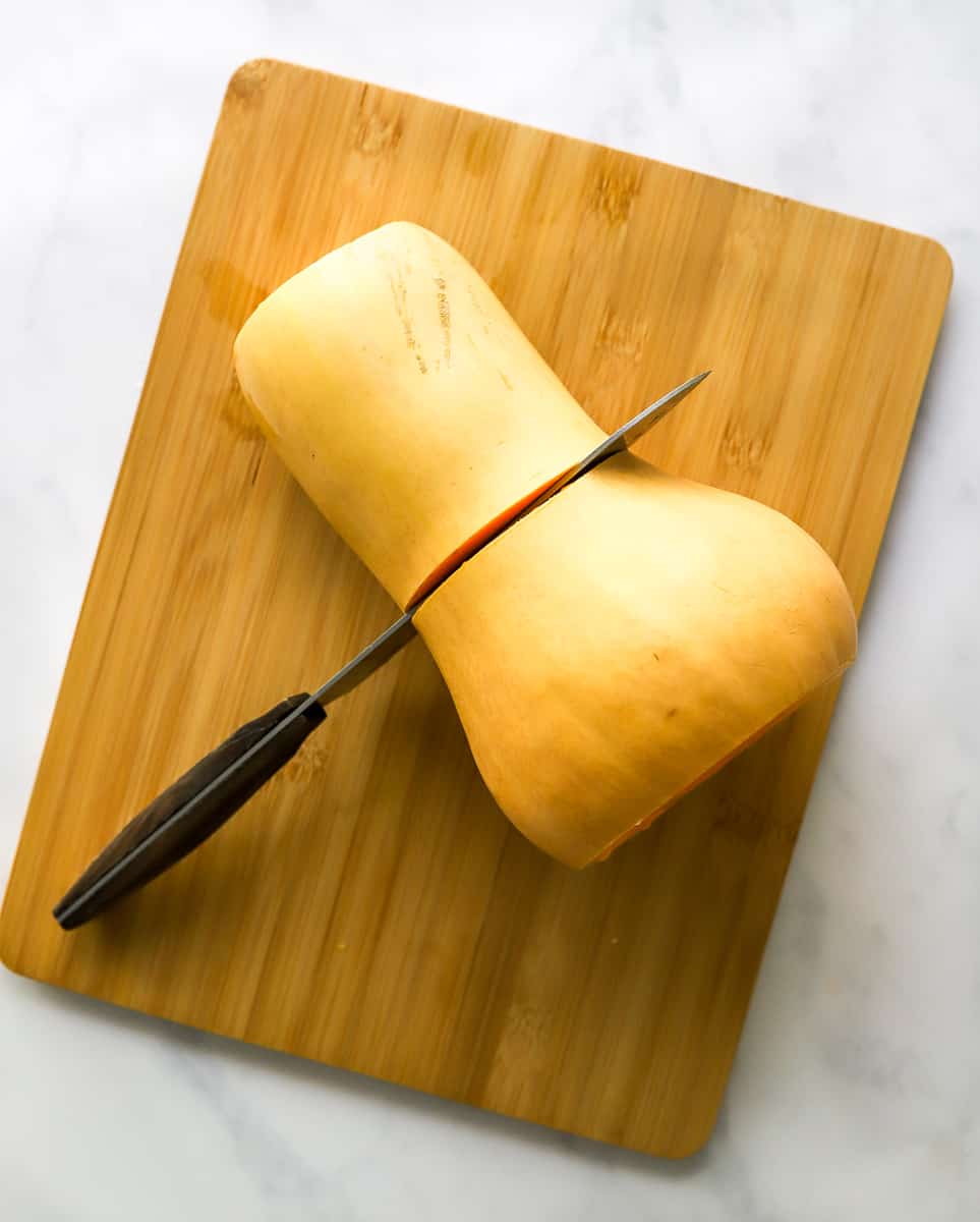 Butternut squash on a cutting board with a knife cutting it in half.