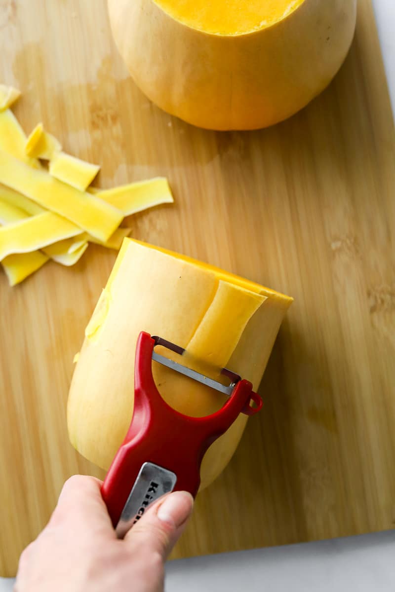 Hand peeling the skin off of a butternut squash with a red vegetable peeler on a wood cutting board.