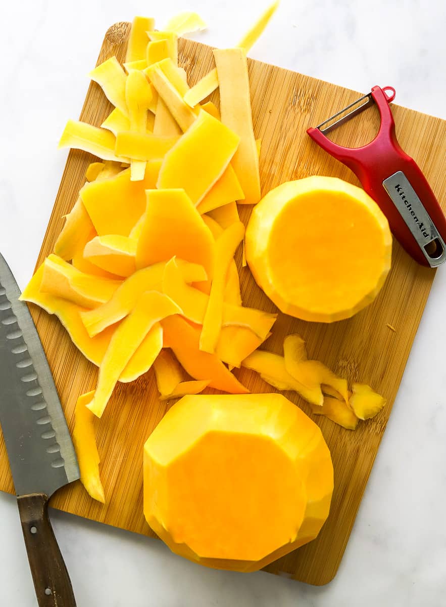 Two halves of squash with peels on a cutting board with a knife and a red vegetable peeler on the cutting board next to it.