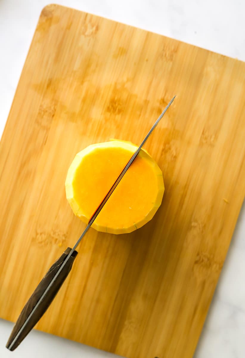 Knife cutting a peeled squash in half that is on a wood board.