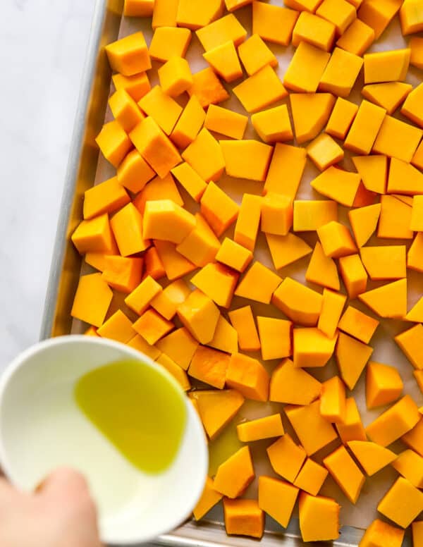 Cubes of cut squash on a baking sheet with a hand pouring olive oil onto the squash.
