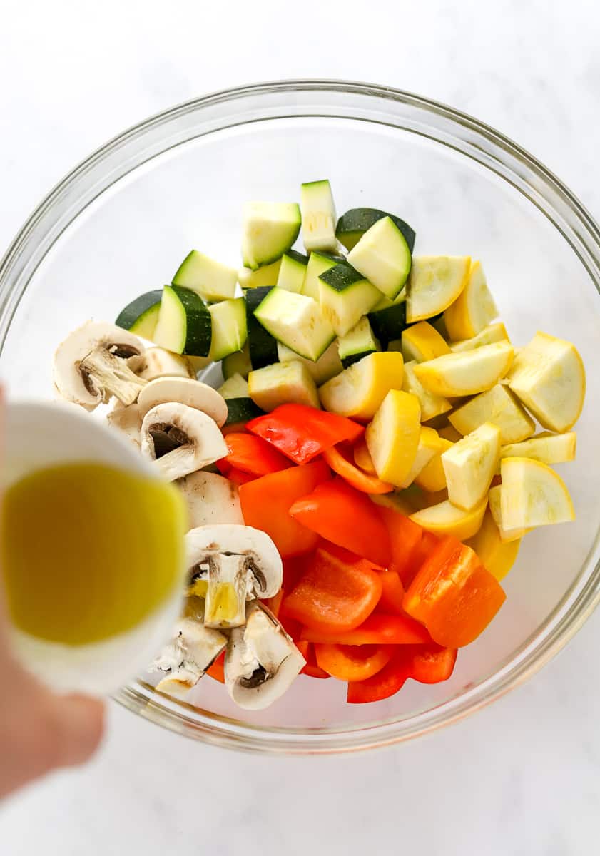 Chopped zucchini, yellow squash, mushrooms, and bell pepper in a glass bowl with oil being poured onto it.