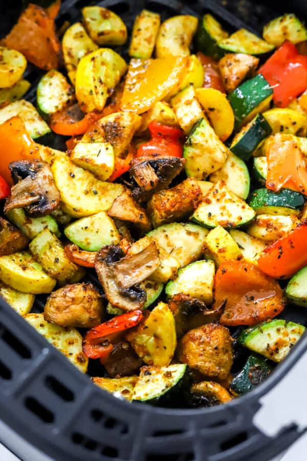 Chopped cooked, seasoned zucchini, mushrooms, bell pepper, and squash in a black, square air fryer basket.