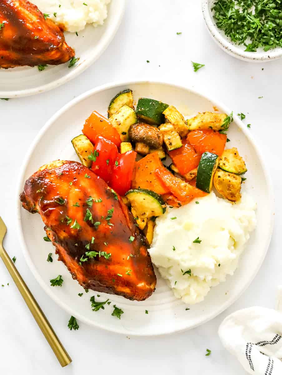 BBQ chicken breast on a white plate with mashed potatoes and roasted veggies next to it with another plate of it behind it and a gold fork next to it.