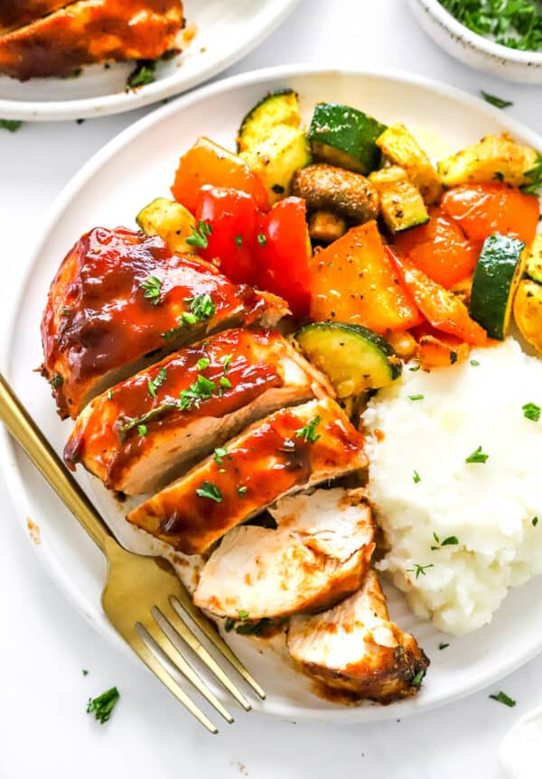 Sliced bbq covered cooked chicken breast on a plate with mashed white potatoes and a medley of cooked veggies with a gold fork on the plate and another plate of chicken behind it.