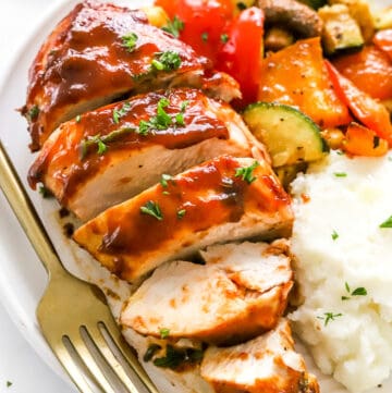 Sliced air fryer chicken breast slathered in bbq sauce, topped with chopped green herbs on a plate with cooked squash and peppers and mashed potatoes with a fork next to the chicken.