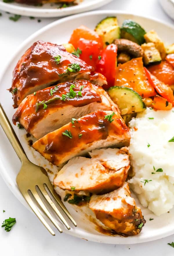 Sliced air fryer chicken breast slathered in bbq sauce, topped with chopped green herbs on a plate with cooked squash and peppers and mashed potatoes with a fork next to the chicken.