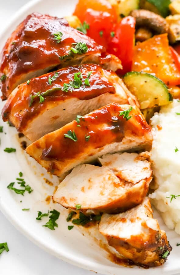 Large piece of air fryer bbq chicken sliced on a plate with potatoes and veggies next to it.