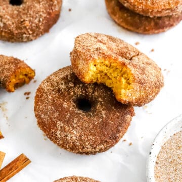 A pumpkin bake donut covered in cinnamon sugar with another donut with a bite out of it on top of it and more donuts around it.