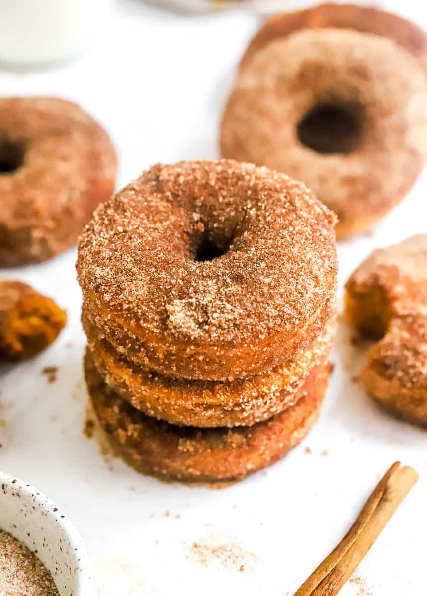 Stack of 3 cinnamon sugar coated pumpkin donuts with ore donuts behind them an a bowl od cinnamon sugar in front of them.