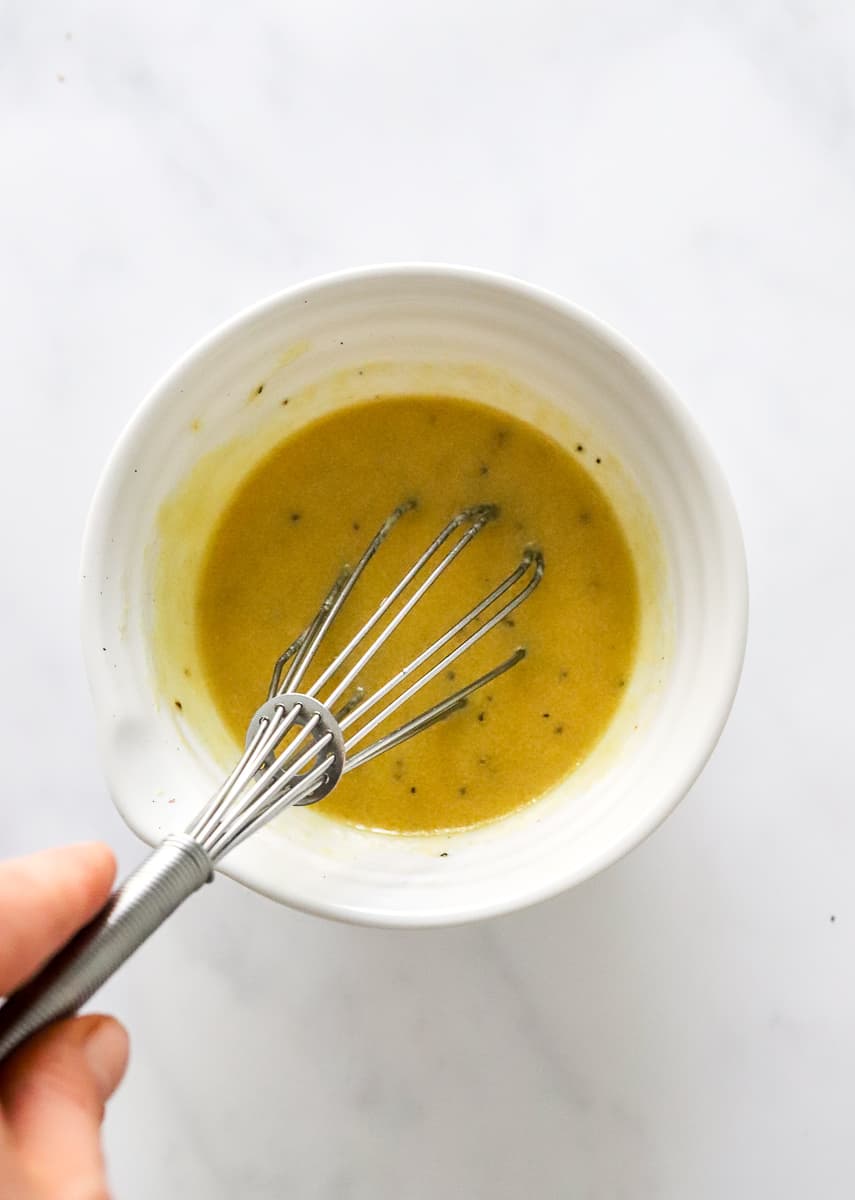 Hand mixing mustard dressing in a white bowl with a metal whisk.