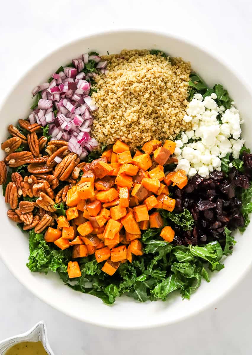 Large round bowl filled with chopped kale, quinoa, diced red onion, crumbles goat cheese, pecans, dried cranberries, and roasted sweet potatoes.