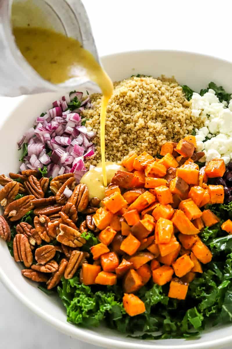 pouring mustard dressing over kale salad with sweet potatoes, pecans, red onion, quinoa, and goat cheese.