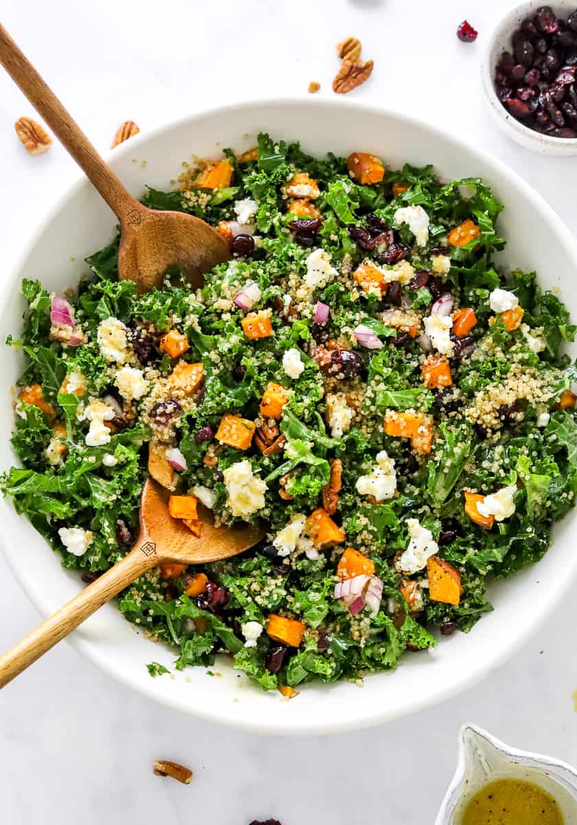 Mixed kale salad in a large white bowl topped with roasted sweet potatoes, diced red onions crimped cheese and nuts.