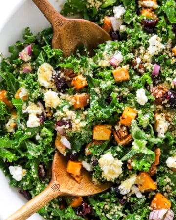 Kale and roasted sweet potato salad in a large salad bowl topped with cheese and cranberries with wooden serving spoons in the bowl.