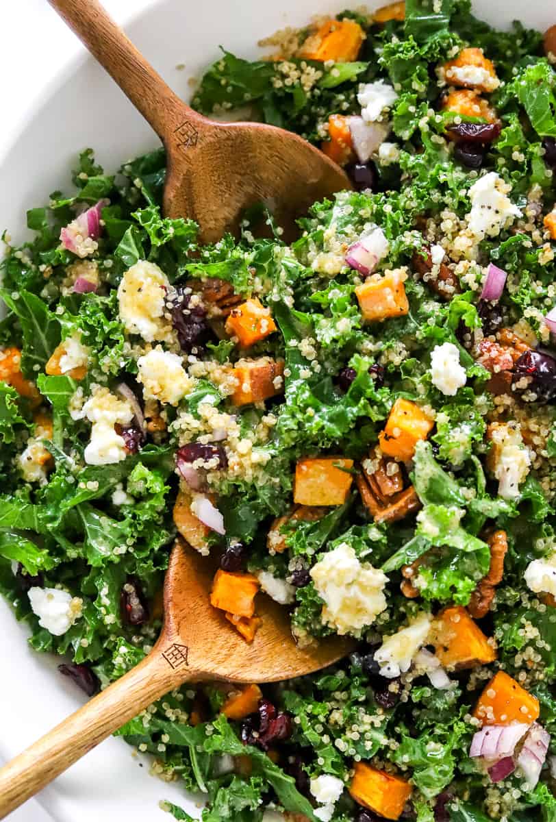 Kale and roasted sweet potato salad in a large salad bowl topped with cheese and cranberries with wooden serving spoons in the bowl. 
