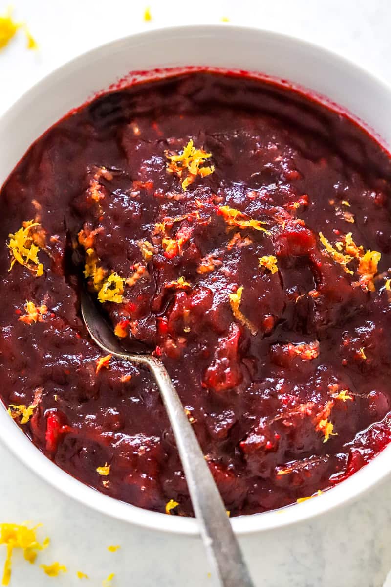 Orange cranberry sauce in a white bowl with orange zest on top of it with a spoon in it. 