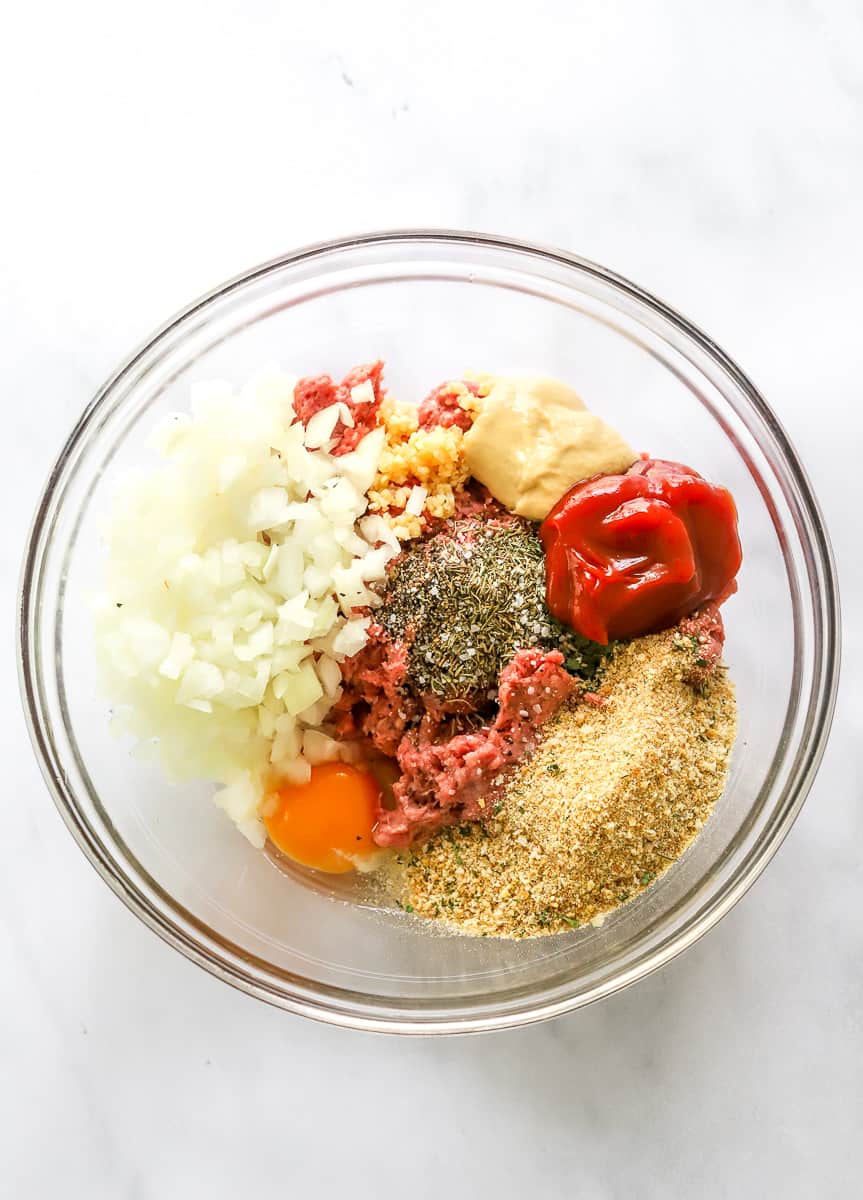 Diced onion, raw beef, egg, ketchup, and breadcrumbs in a glass bowl.