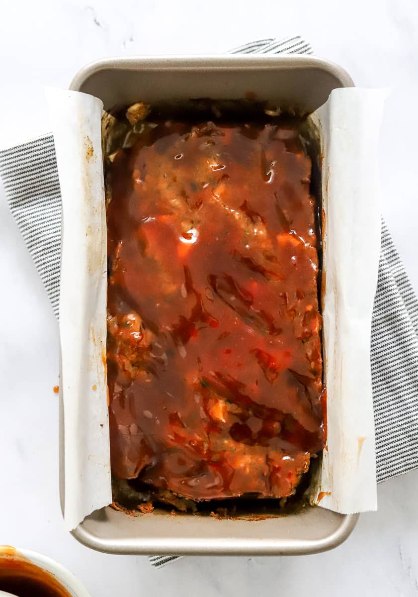 Cooked meatloaf in a loaf pan with tomato glaze on top of it with more of the glaze in a bowl in front of it.