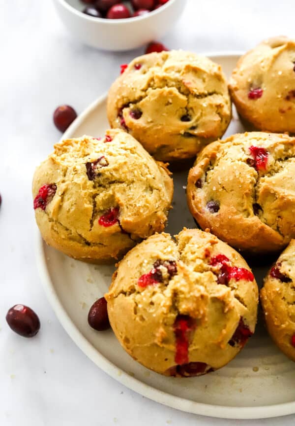 Plate filled with baked muffins with cranberries in them with a few cranberries scattered around them and some behind them.