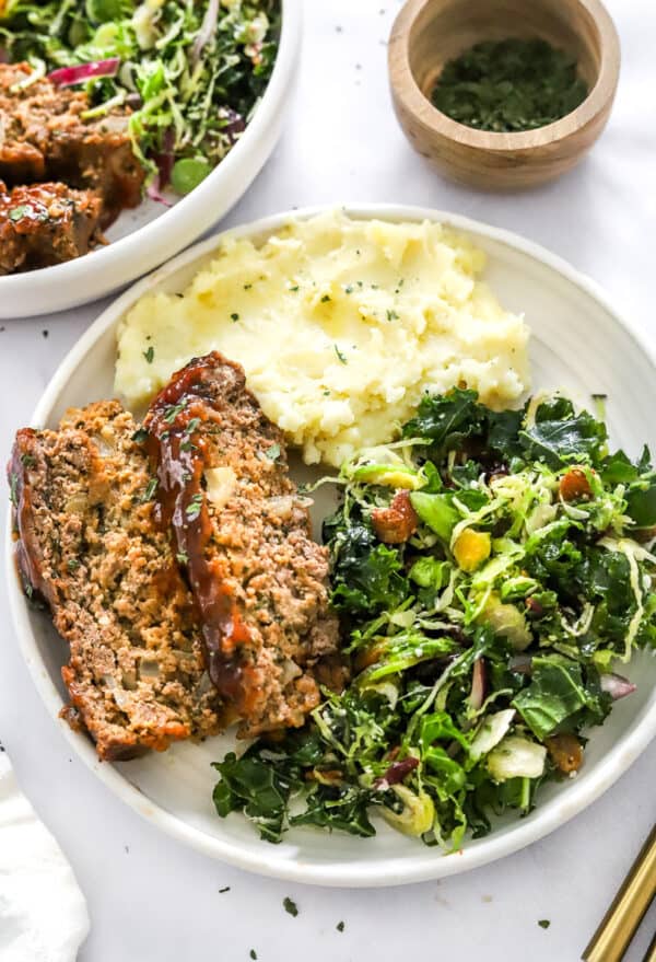 Plate of meatloaf with kale salad ad mashed potatoes on the plate with it and another plate of it behind it.