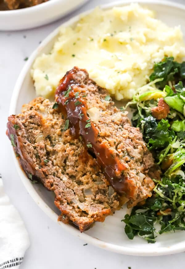 Sliced meatloaf with tomato glaze on it on a white plate with mashed potatoes behind it and a Brussel sprout kale salad next to it on the plate.