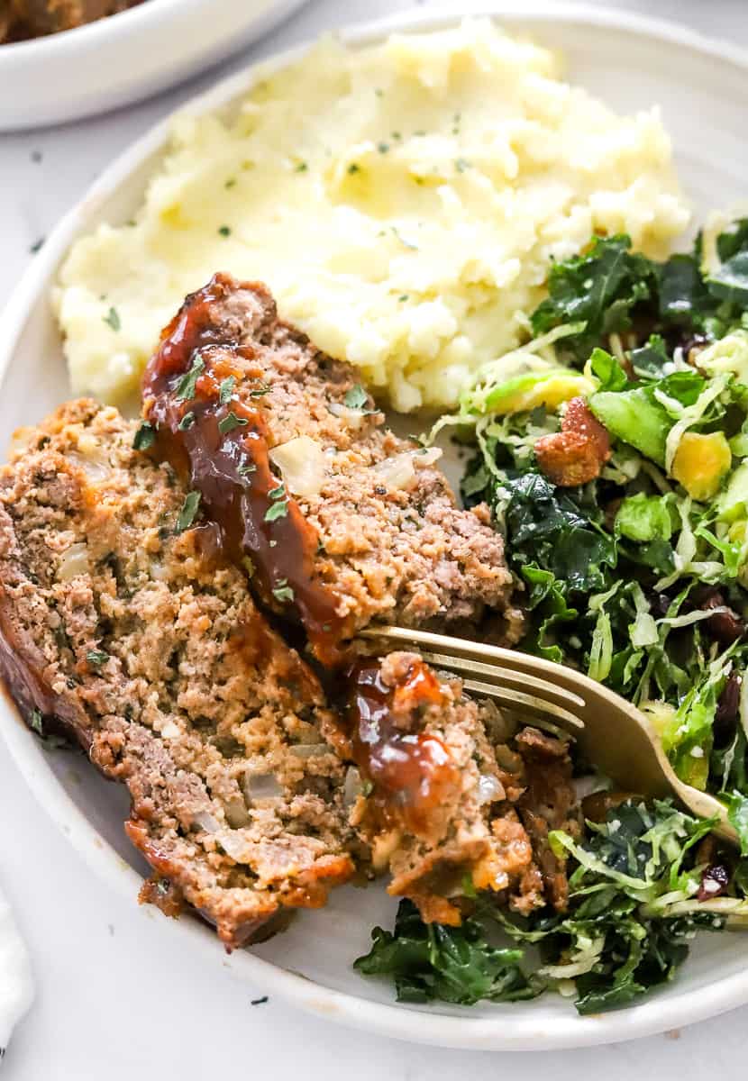 Cooked, sliced air fryer meatloaf on a plate with a fork cutting through it with mashed potatoes and green kale salad next to it on the plate.