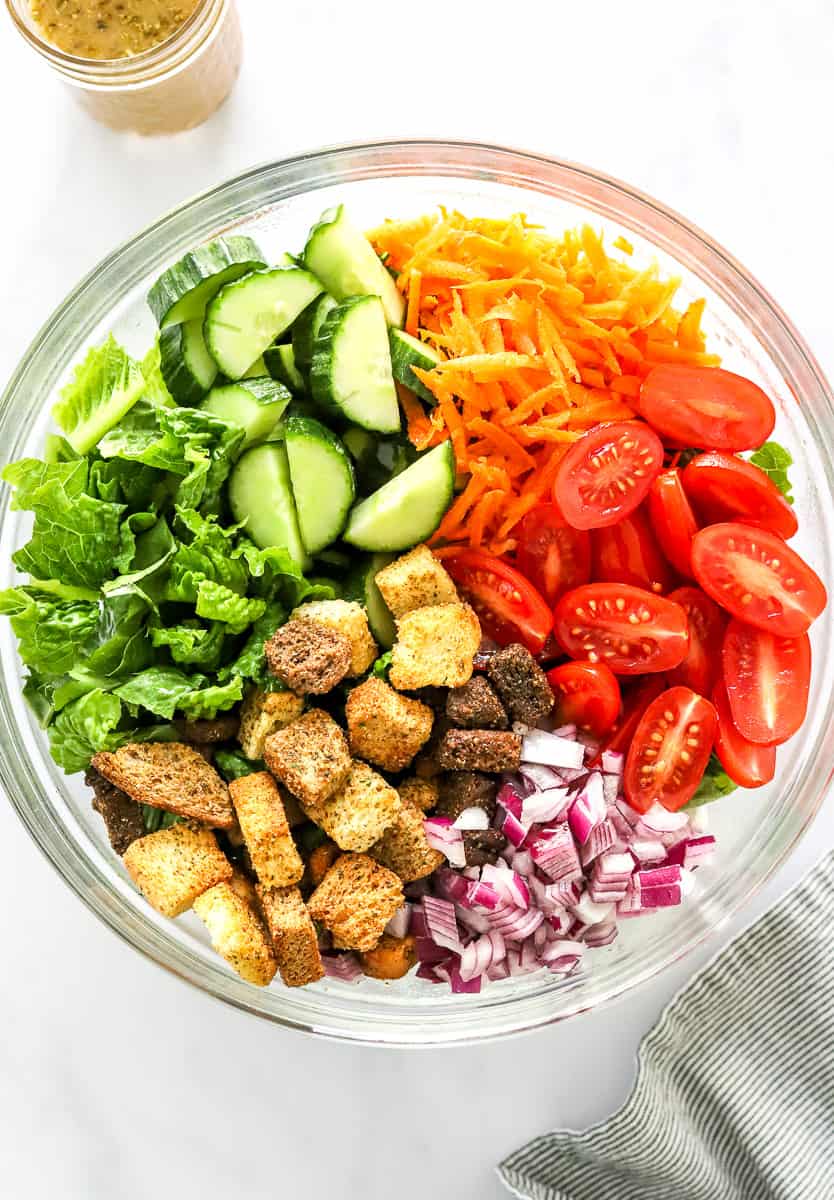 Chopped lettuce, cucumber, tomato, shredded carrots, diced red onion, and croutons in a large glass salad bowl. 