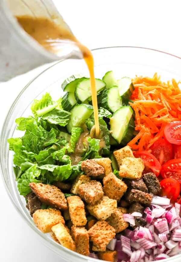 Chopped lettuce, carrots, cucumber, onion and croutons in a large bowl with salad dressing being poured over it.