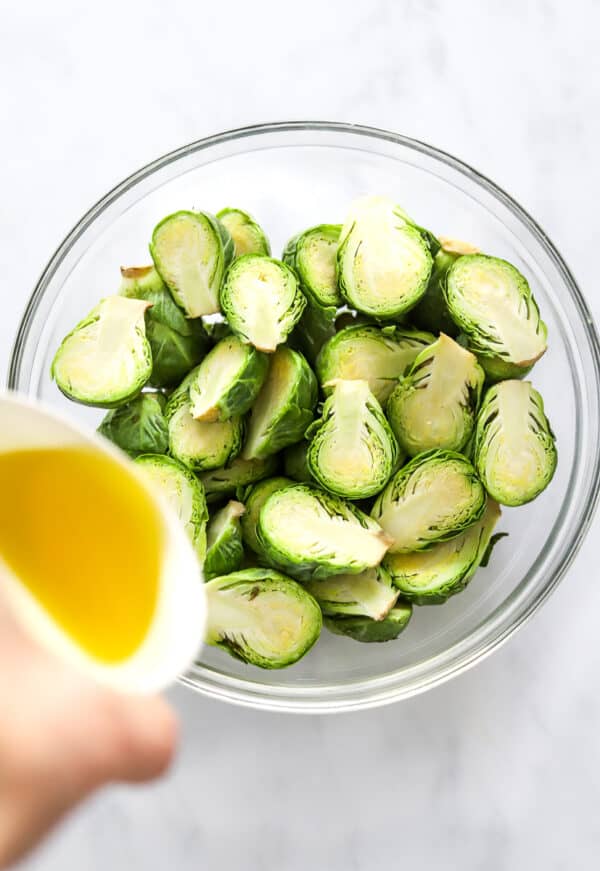 Sliced Brussels sprouts in a glass bowl with olive oil being poured over them.