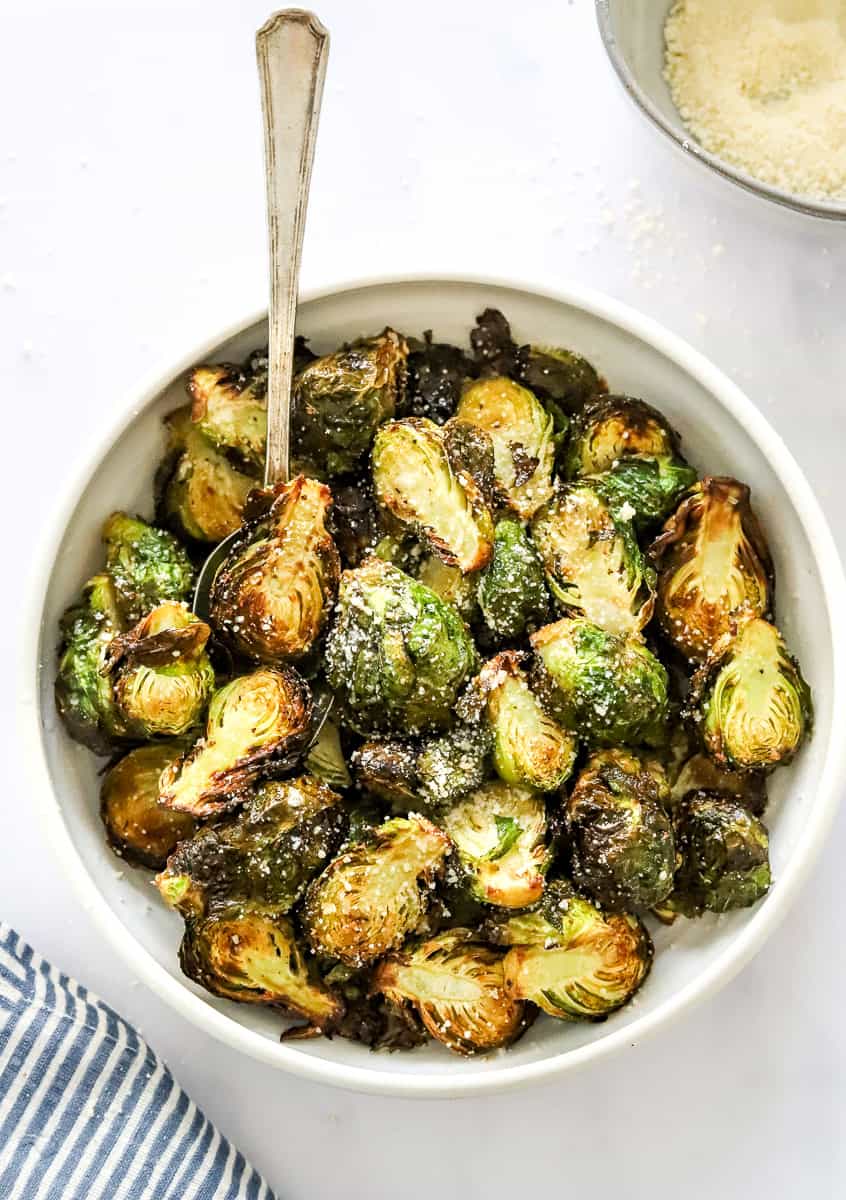 Bowl of golden charred Brussels sprouts with a spoon in the bowl and a bowl of grate parmesan cheese behind them.