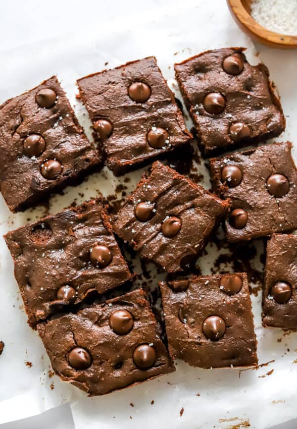 Cut cooked dark chocolate brownies on top of white parchment paper with a wooden bowl of sea salt behind it.