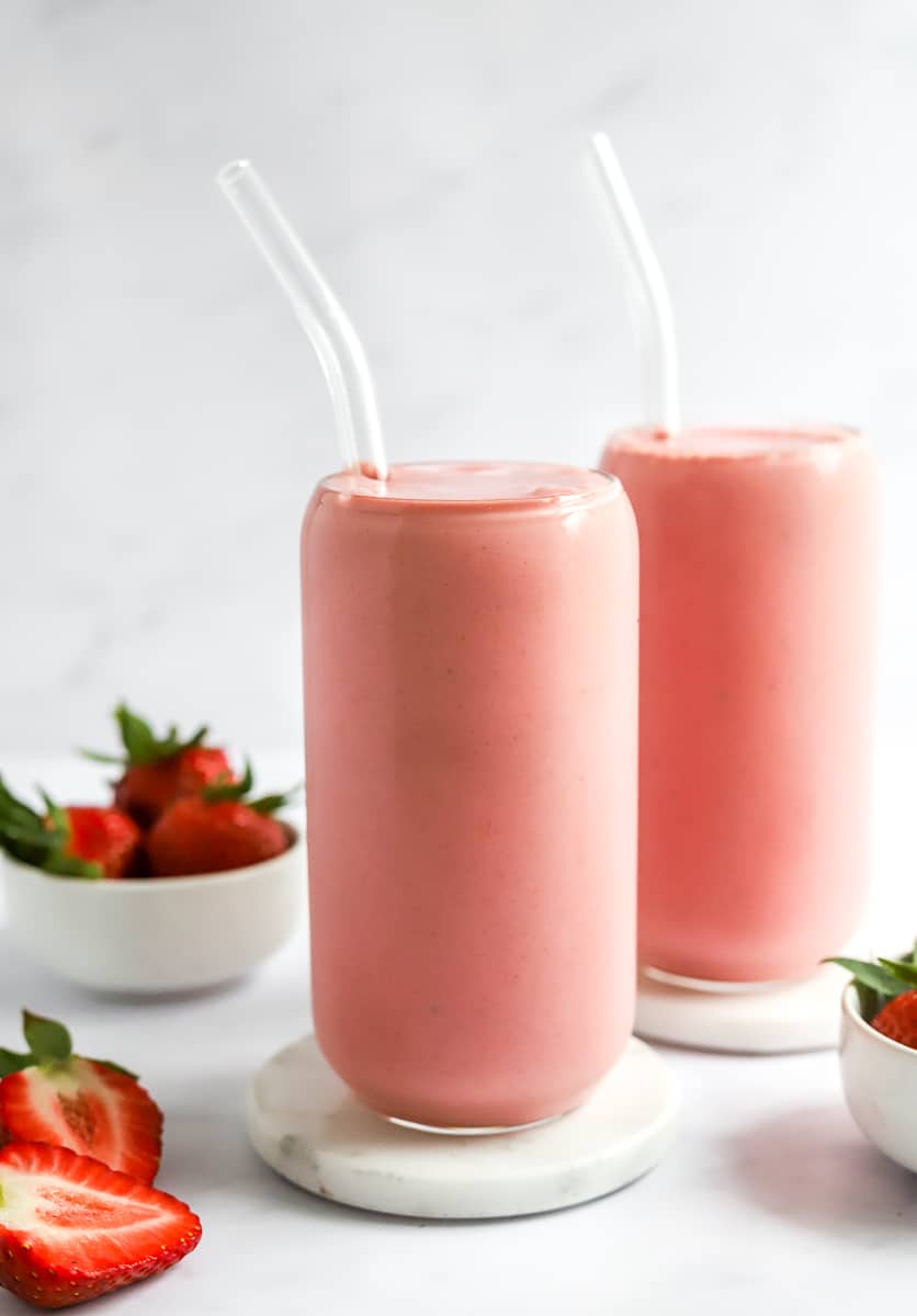 Two glasses filled with strawberry banana smoothie with glass straws in the glasses and strawberries behind it and a cut strawberry in front of it.