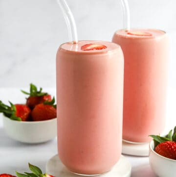 Two tall pink smoothies in glasses with glass straws with strawberries around the.