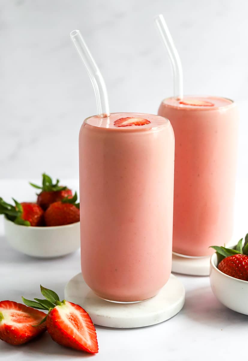 Two tall pink smoothies in glasses with glass straws with strawberries around the.