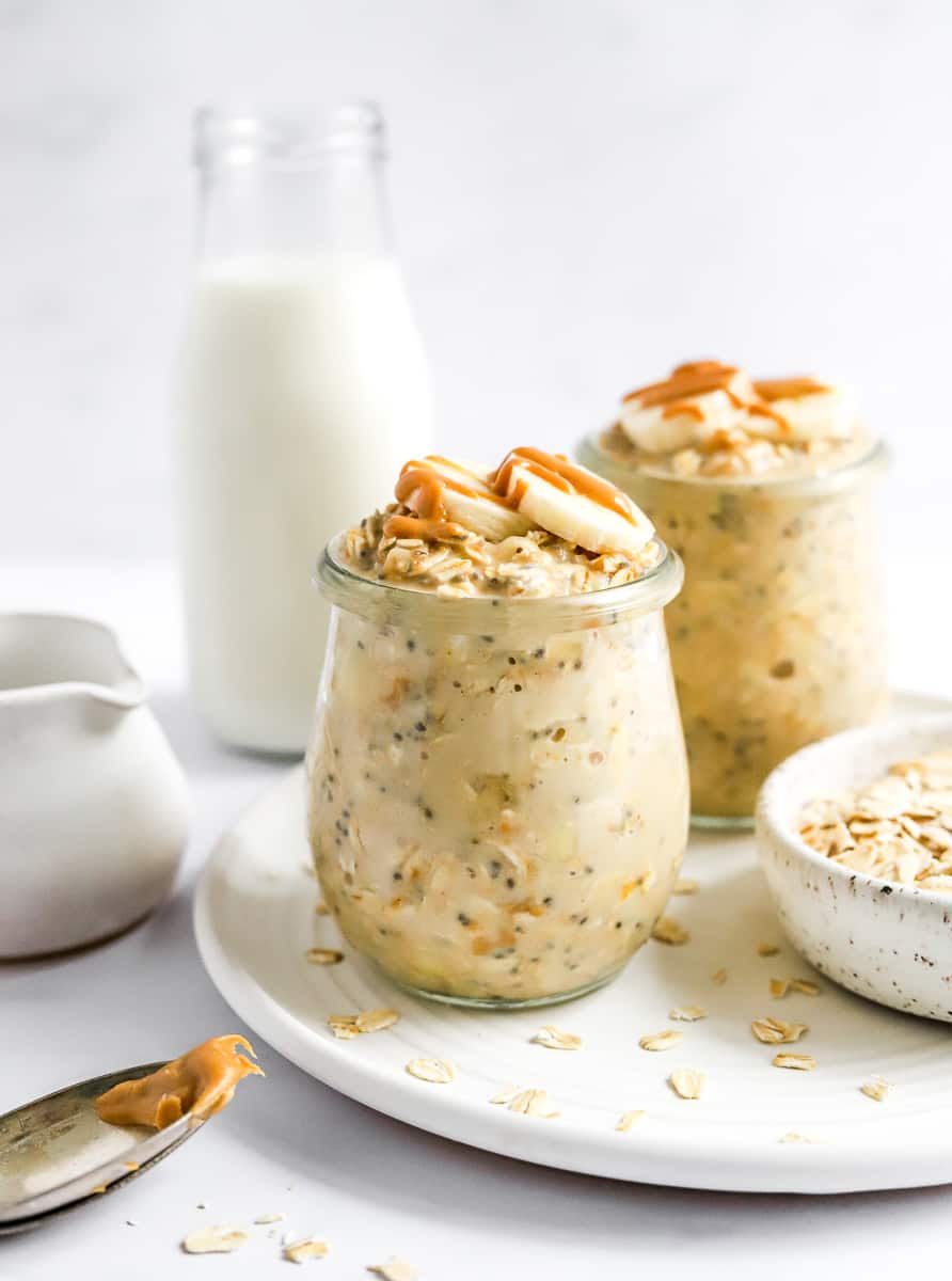 Two glass jars filled with peanut butter overnight oats topped with sliced banana with a jar of milk behind them and a bowl of raw oats next to them. 