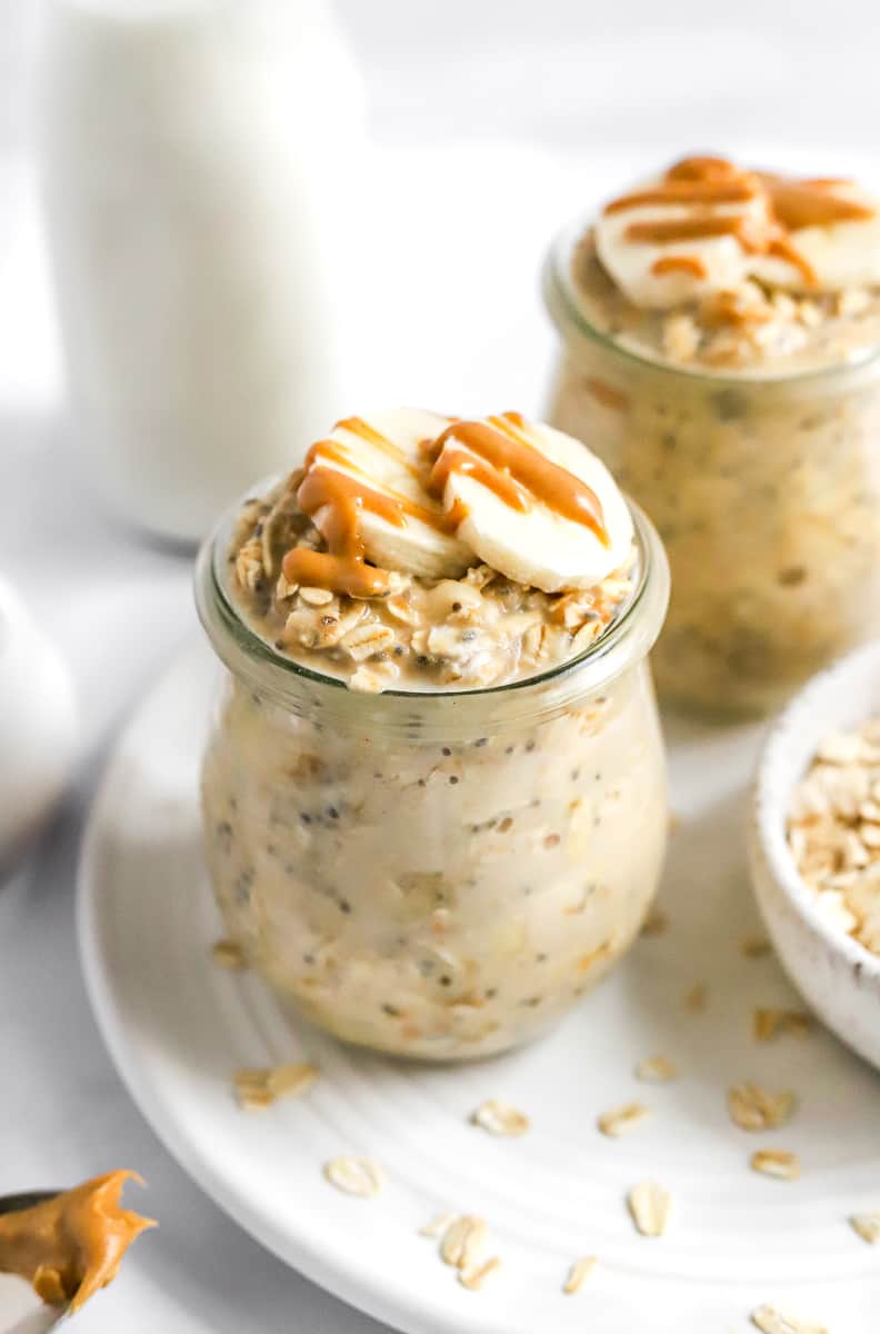 Glass jar filled with creamy oats topped with sliced banana and drizzle of peanut butter with another jar of oats behind it. 