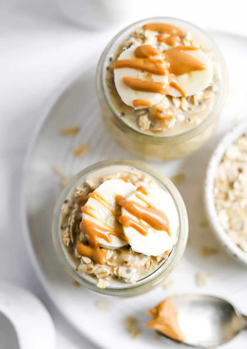 Two glass jars filed with oatmeal topped with slices of banana and a drizzle of peanut butter on a plate. 