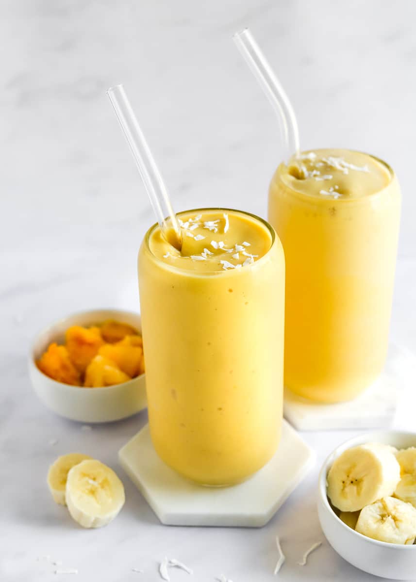 Two glasses filled with a mango banana smoothie with glass straws in them and shredded coconut on top with a dish of mango behind it and a dish of sliced banana in front of it. 