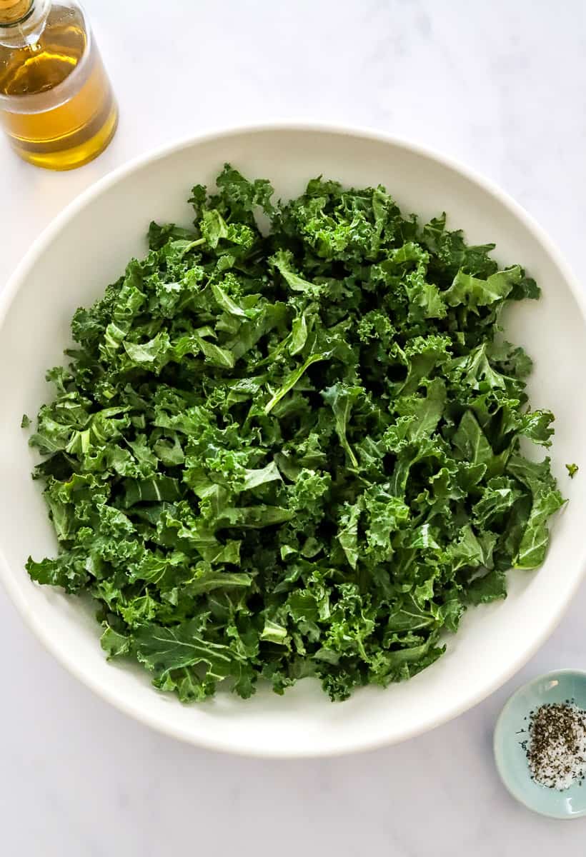 Chopped green kale in a white serving bowl with olive oil behind it and a bowl of salt and pepper in front of it.