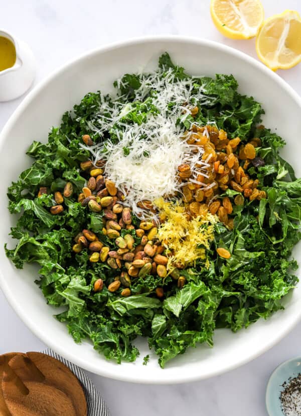 Chopped kale salad in a bowl with nuts, shredded cheese, raisins and lemon zest in the middle of it with sliced lemon behind it and wooden serving spoons in front of it.