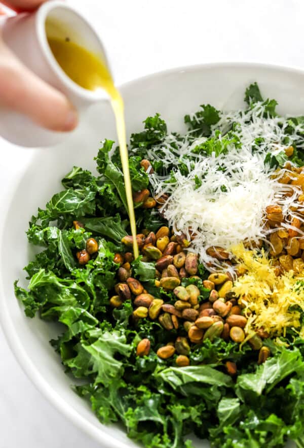 Hand pouring lemon dressing over kale salad in a white bowl.