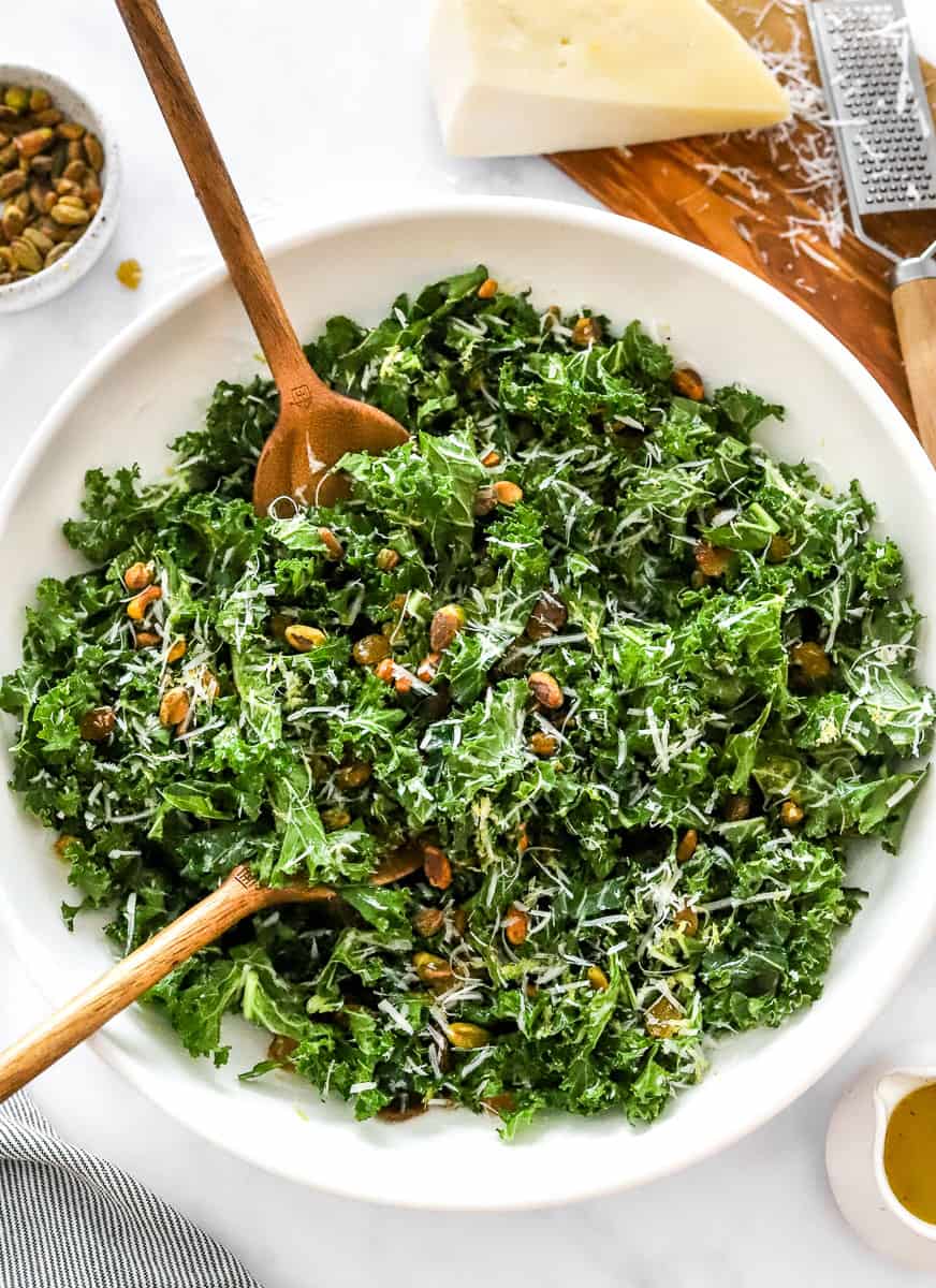 Mixed kale with pistachios, shredded cheese and raisins in a white salad bowl with wooden spoons in the bowl with a block of parmesan on a board and a bowl of pistachios behind it. 