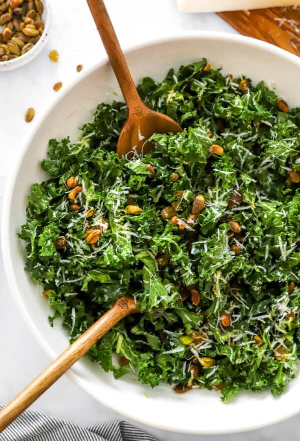 Mixe kale salad in a white salad bowl with wooden serving spoons in the bowl.